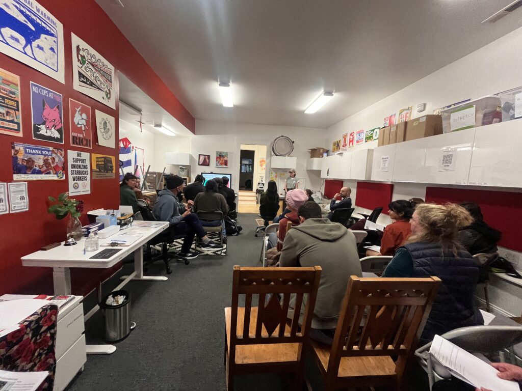 A photo of about 20 people sitting in the DSA SF office during a Know Your Rights training, listening to a speaker at the front of the room. The photo is taken from the back of the room, facing toward the speaker.