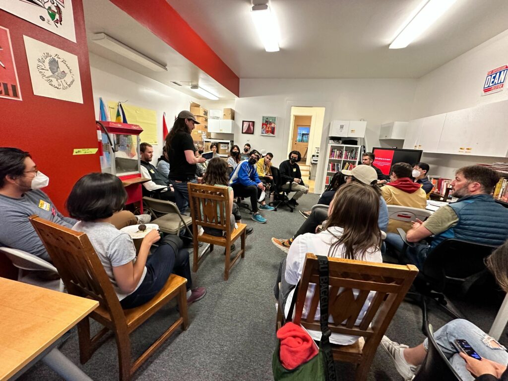 A photo of comrades gathered in the DSA SF office, seated in chairs while one person stands up to speak.