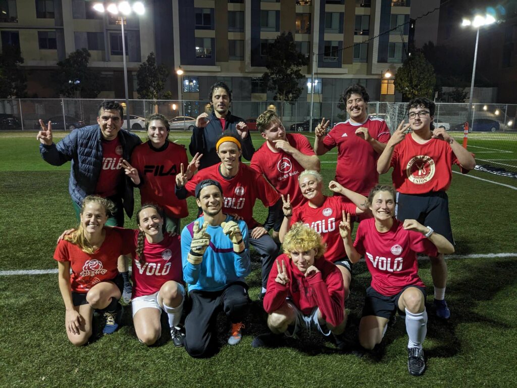 The thirteen members of the DSA SF Orcas soccer club pose together on the soccer pitch, celebrating their win.