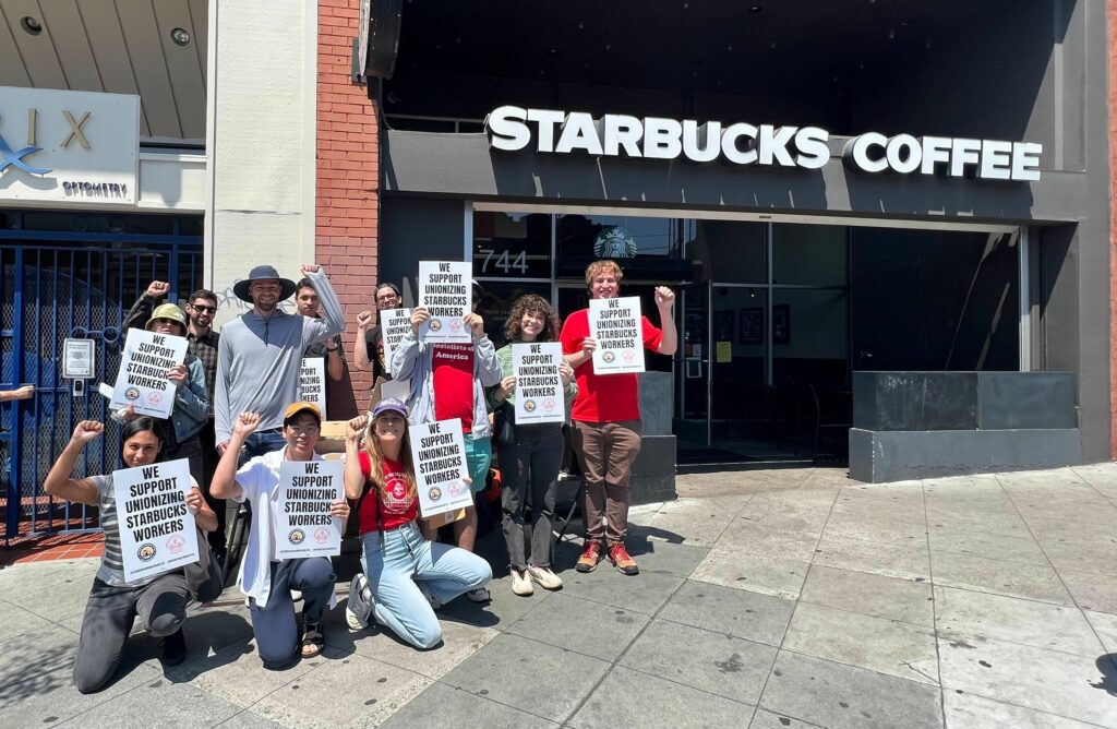 Several DSA members pose for the camera outside the 9th and Irving Starbucks. Everyone's fists are raised, and most are holding posters that say "We Support Unionizing Starbucks Workers."