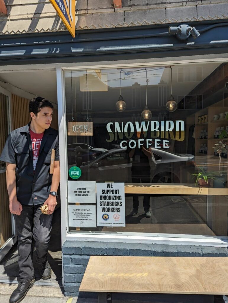 The front door and window of Snowbird Coffee. Someone is standing in the doorway. In the front window, a poster is displayed that reads, "We Support Unionizing Starbucks Workers."