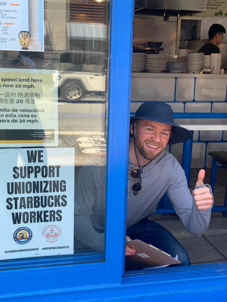 A person sitting inside a restaurant by an open window, smiling and giving a thumbs up to the camera. In the window, there's a poster that says "We Support Unionizing Starbucks Workers."
