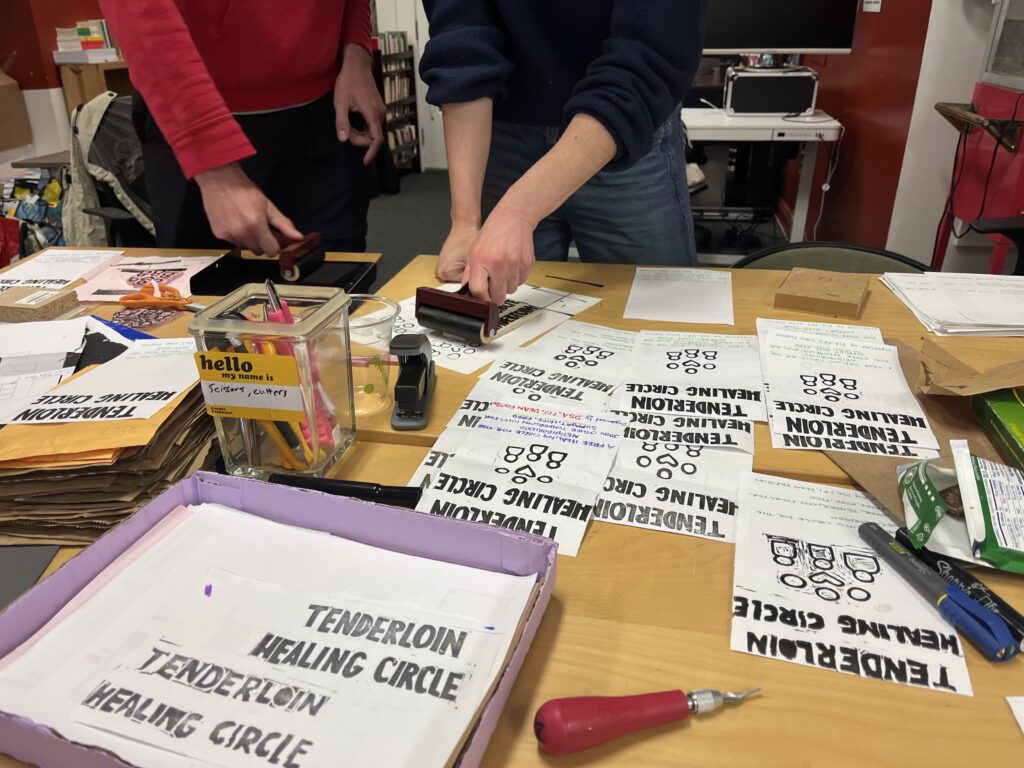 A photo of many flyers for the Tenderloin Healing Circle spread across a table. Two people work together to print another flyer using ink rollers and a hand-carved rubber stamp.