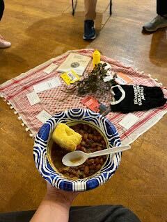 A photo of someone holding a bowl of hearty food. In the background, the edge of a circle of chairs and a red keffiyeh can be seen.