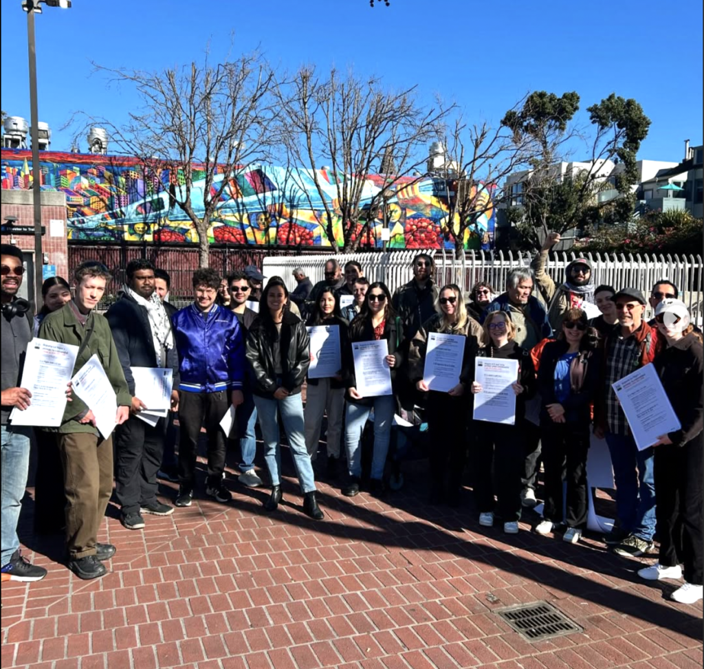 A group of about thirty people, including Supervisor Jackie Fielder, pose for a photo at the 24th St. Mission BART station plaza.