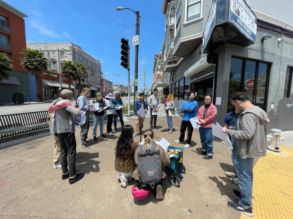 A little over a dozen people stand in a circle on a street corner, holding papers during a Know Your Rights training.