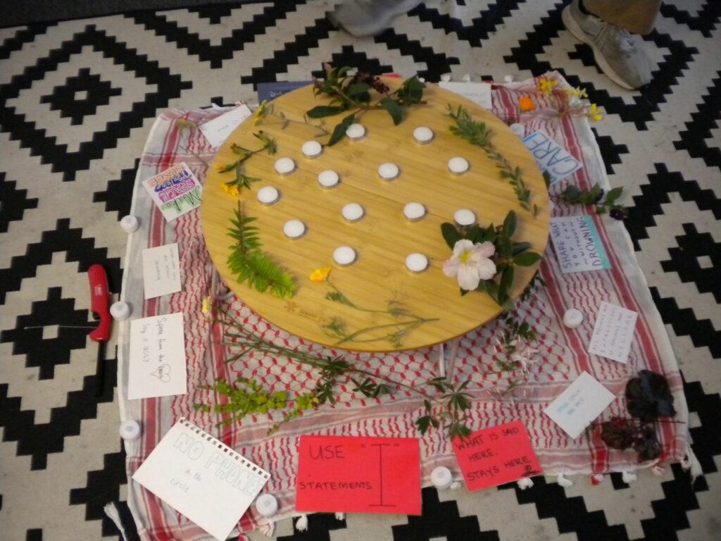 A photo of flowers, plants, candles, notes, and stickers sitting on top of a red keffiyeh.
