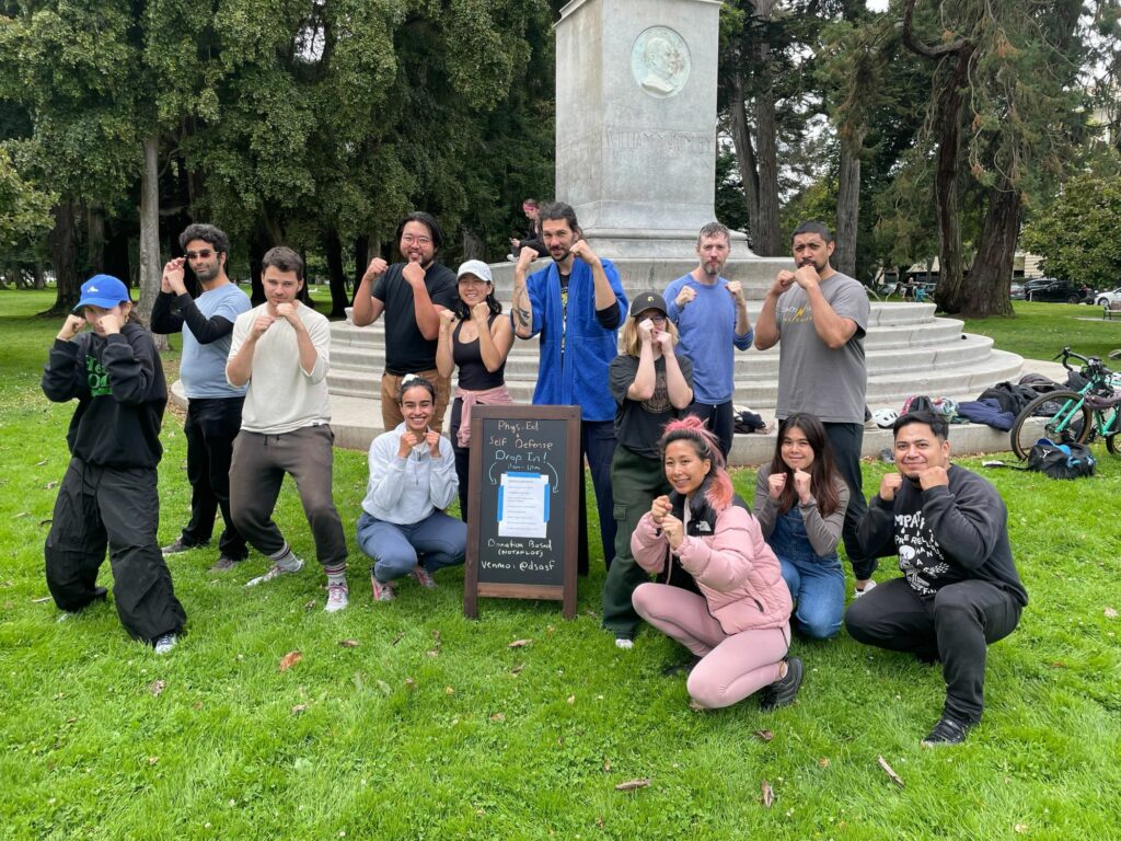 13 people pose outside in a park while smiling with their fists up.