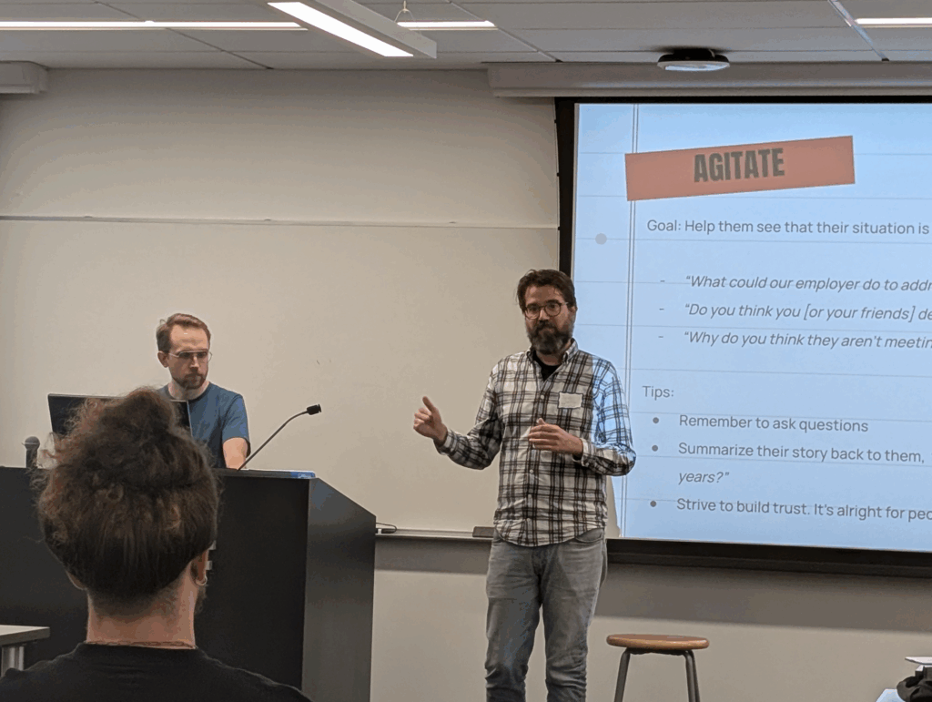Two people at the front of a classroom, one behind a podium, the other gesturing while standing in front of a projected presentation with the word "AGITATE" in bold lettering on the screen. The back of another person facing them can be seen in the foreground.