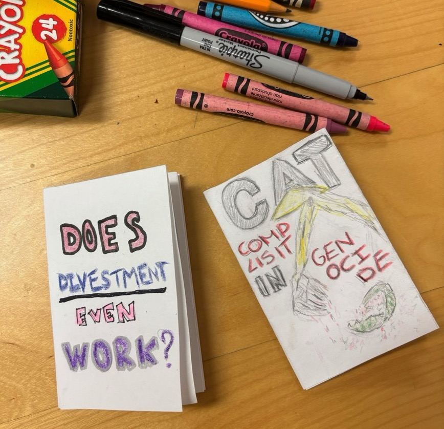 A photo of two zines, some crayons, and a sharpie on a wooden table. The left zine's cover says "DOES DIVESTMENT EVEN WORK?" with the word 'divestment' underlined. The right zine's cover has a drawing of the arm of an excavator. Below it is a watermelon split in two. The excavator bucket is dripping red watermelon juice. Its title reads, "CAT", and below, "COMPLISIT [sic] IN GENOCIDE".